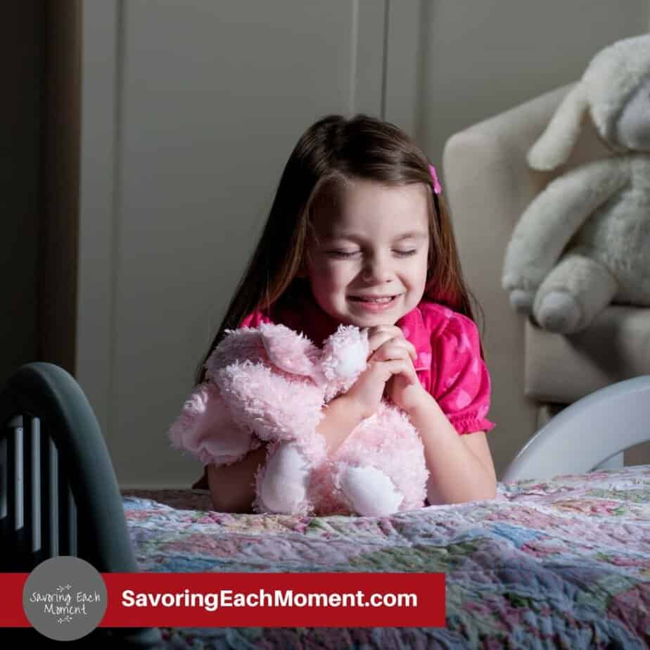 Little girl in pink shirt holding a stuffed animal praying beside her bed at night.
